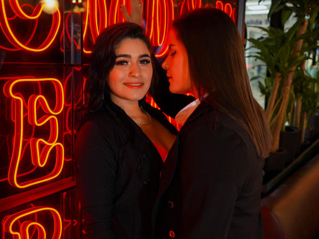 MaraANDSara, avec ses cheveux noirs et son anneau de nez, portant un cardigan noir, se tient à côté de Sara, qui a de longs cheveux bruns, dans une pièce faiblement éclairée avec des lumières rouges néon..