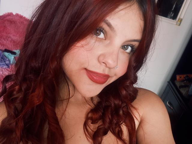 Sophie Hill, avec ses cheveux roux bouclés, son anneau de nez et son rouge à lèvres rouge, sourit à l'appareil photo, montrant ses épaules dénudées, avec un look à la fois mignon et sexy..