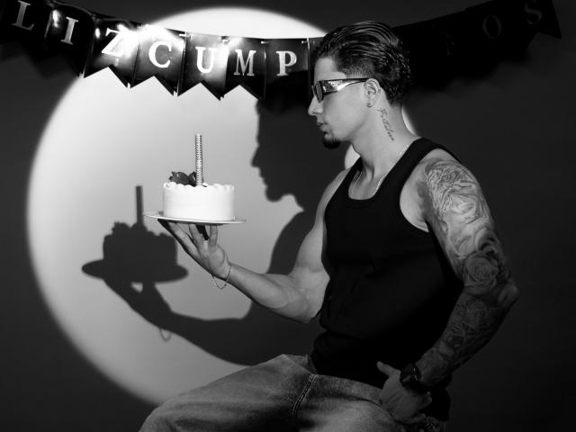 Zwart-wit foto van CamiloAlva met tatoeages, die een tanktop en jeans draagt, en een taart met een kaars vasthoudt. Er is een schaduw op de muur. Hij draagt een zonnebril en kijkt naar de zijkant..