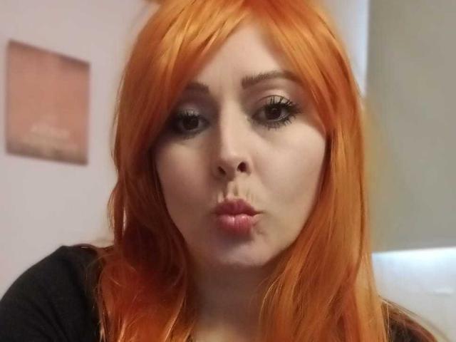 HesteSiaX has bright orange hair, puffy lips in a kissing pose, and thick black eyelashes, wearing a black top, with pale skin and a slightly surprised expression.