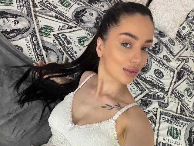ArielGemme with long black hair, white lace camisole, small tattoo on right collarbone, lying on money-filled bed, looking at camera with slightly parted pink lips.