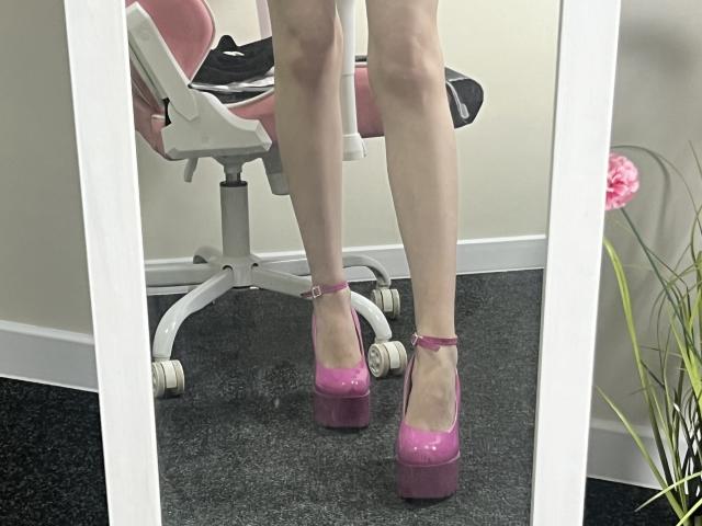 Valnes' legs in pink platform heels, standing in front of a mirror. She's wearing a pink and black chair, white wall, and a pink flower in the background.