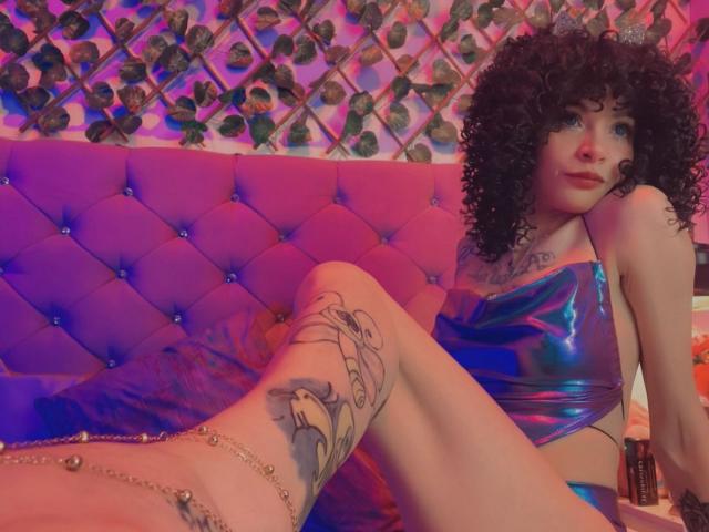 LaurenEvie69 with curly black hair, wearing shiny purple-blue bikini top, sitting on purple tufted couch, showing tattooed leg, ankle chain, and purple-blue light.