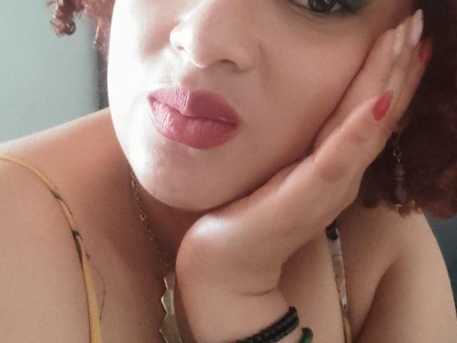 VioletaSwitch with red lipstick, curly brown hair, green eyeshadow, pale skin, hand on cheek, red nail polish, yellow and black patterned top, necklace, and black bracelet.