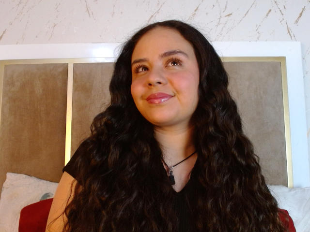 ArielleWilde with long, wavy black hair, wearing a black shirt, smiling at the camera, showing a small nose and a cute, slightly chubby face with pink lips.