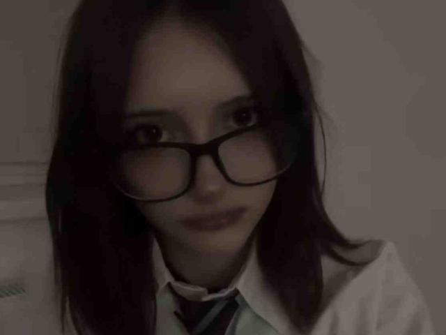 DelorisBueno with dark brown hair, wearing black glasses and a white shirt with a striped tie, smiling slightly. She has a cute face and is looking at the camera.