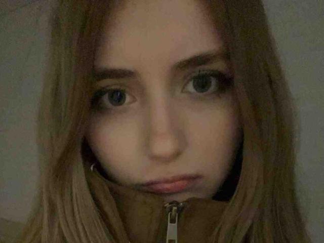 DelorisBueno's face, with her blue eyes looking slightly to the side, has long brown hair and a serious expression, wearing a brown zip-up, in a dimly lit photo.