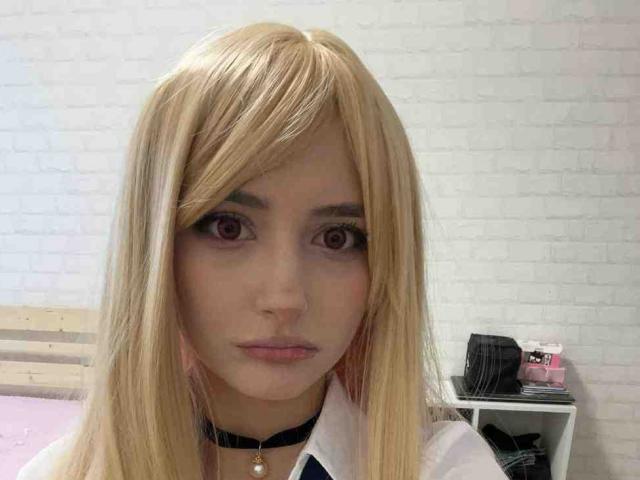 DelorisBueno with blonde wig, pink eyes, pale skin, black choker, and white shirt, looking straight at camera, bedroom background, light pink bed, white brick wall.