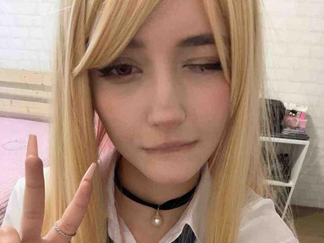 DelorisBueno with blonde hair, winking, making a peace sign, wearing a black choker with a pearl, in a white-walled room. Her pink eyes and subtle smile are visible.