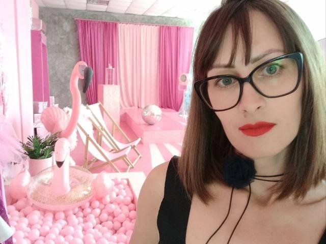 MiraMixi with red lipstick, black glasses, and a black choker, standing in a pink room with a flamingo decoration, pink bubble floor, and pink curtains.