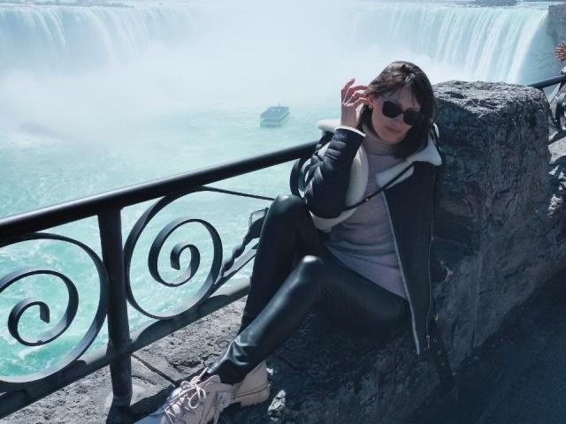 MiraMixi sits on a rock, wearing a black jacket, white hoodie, black leather pants, and white sneakers, with sunglasses on, near a waterfall, touching her hair.
