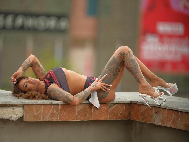 Tara Elliot, con tatuajes, acostada sobre un borde de ladrillo, vistiendo un bikini morado y rojo, tacones plateados altos, piernas levantadas, mano en la cabeza, mirando a la cámara, sonriendo..