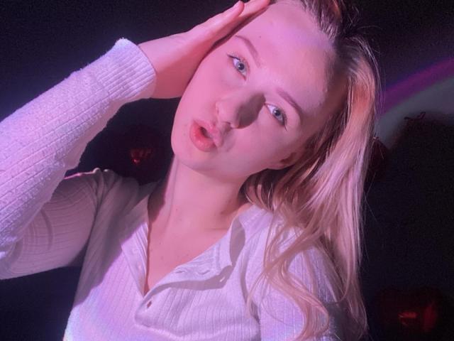FlyFreedom has blonde hair, blue eyes, and pouty pink lips, wearing a white ribbed shirt, hand on head, in a dimly lit, pink-hued photo. Her tits are small.