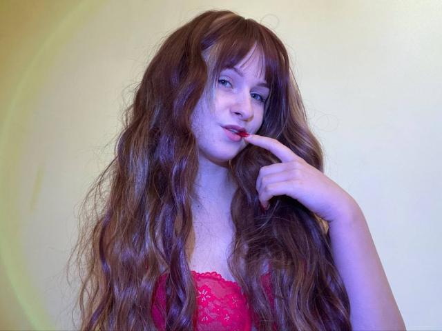FlyFreedom with long, wavy brown hair, wearing red lace lingerie, has a finger to her slightly open mouth, showing a seductive look. Her tits are visible under the lace.