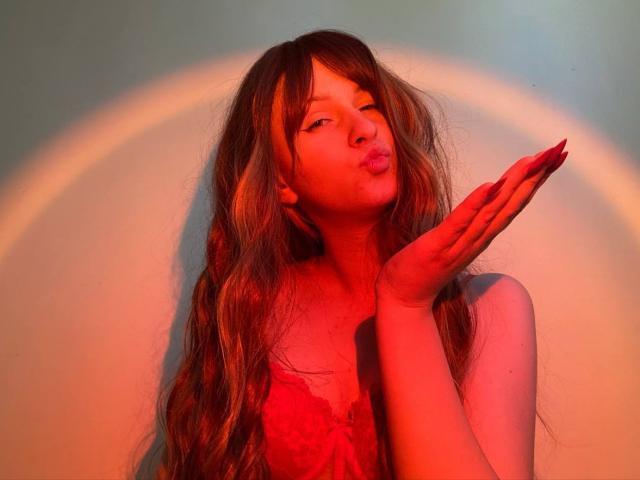 FlyFreedom with long, wavy brown hair, pouty lips, and red nails, hands up, wearing a red lace top, illuminated by warm orange-red light.