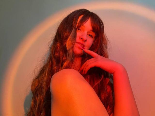 FlyFreedom with long, wavy brown hair, red-tinted skin, and a finger touching her slightly parted lips. She's topless, with a soft, seductive expression and a rainbow shadow.