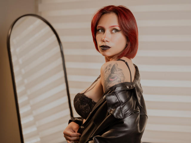 LilithDeveraux with red hair, black lipstick, leather jacket, and lace bra, showing off her tattooed shoulder and looking over her shoulder at the mirror. Her tits are visible.
