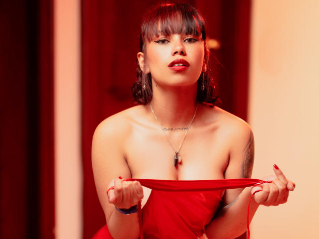 Lilith Deveraux, com um vestido vermelho puxado para baixo, revelando seus seios, usando um colar, batom vermelho e uma pulseira azul, está em pé em um quarto com iluminação vermelha, olhando sedutoramente para a câmera..