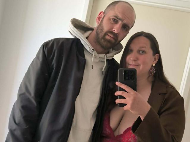 CoupleLovve69 avec un homme chauve, barbu, portant une veste noire et un sweat à capuche, debout à côté d'une femme aux cheveux bruns, vêtue d'un manteau marron et d'un soutien-gorge en dentelle rouge, prenant un selfie..