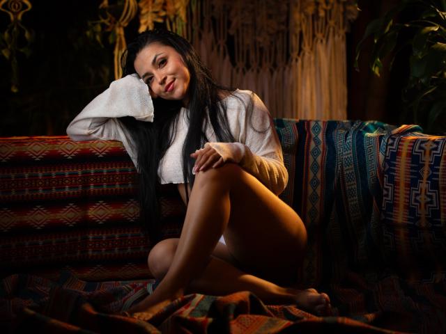 HikariChloe, with black hair, white shirt, and no pants, sits on a colorful, patterned couch, smiling, legs crossed, arm resting on her head, showing her bare ass.