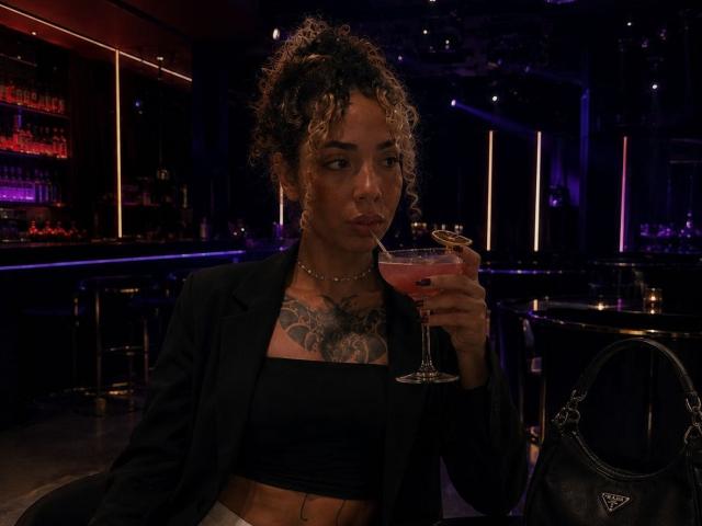 SilvanaXOwen with curly hair, black tube top, black blazer, holding a pink cocktail, tattoo on chest, black handbag on bar, dim lighting, looking serious.