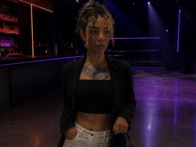 SilvanaXOwen with curly hair, black crop top, black blazer, white pants, chest tattoo, hands in pockets, purple lights, serious expression, standing in dimly lit bar.