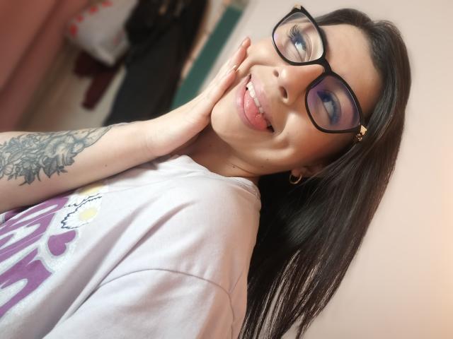 AnntonellaWatson with black glasses, sticking out tongue, hand on cheek, tattoo on arm, hoop earrings, long black hair, purple shirt, smiling, cute, attractive, sexy.