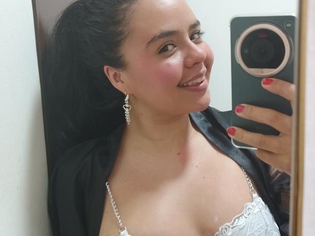 ArielleWilde with dark hair in a ponytail, wearing a white lace bra, black shawl, and heart earrings, smiling at a mirror selfie, holding a green phone with red nails.
