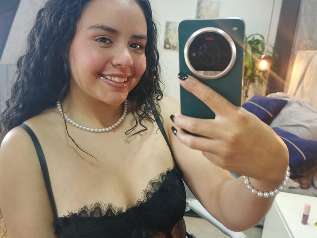 ArielleWilde with curly black hair, smiling, wearing a black lace bra, pearl necklace, and bracelet, taking a selfie with a green phone, showing her tits and black nails.