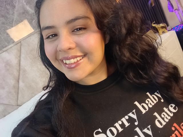 ArielleWilde has a cute smile with glossy pink lips, curly black hair, and a cute nose. She's wearing a black shirt, looking sexy with her tits partially visible.