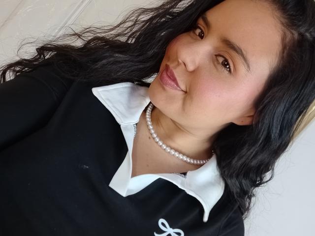 ArielleWilde with wavy black hair, brown eyes, and pink lipstick, wearing a black shirt with a white collar, and a pearl necklace, smiling slightly.