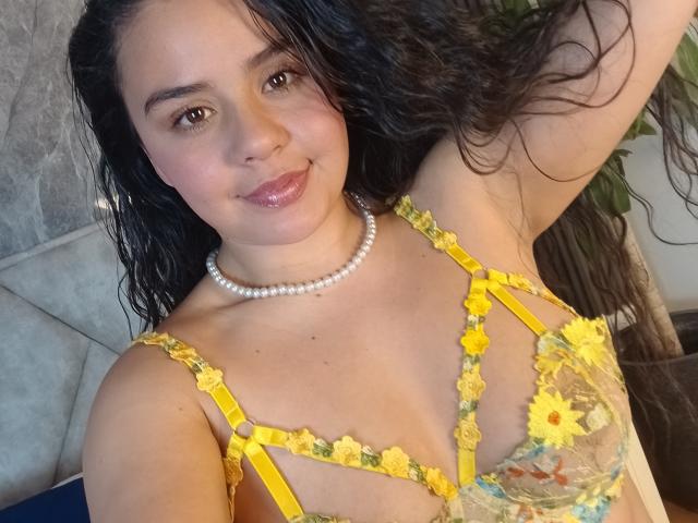 ArielleWilde with wavy black hair, smiling, wearing a yellow floral lace bra, pearl necklace, and showing her small tits. Her skin is tan, and she's looking at the camera.