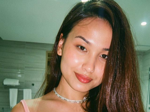 LuccyGrey with brown hair, medium skin tone, and red lipstick, wearing a pink top and silver necklace, smiling softly. Her tits are small, and she has a cute nose.