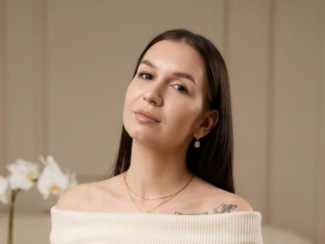 TonyaBartron has long brown hair, a subtle smile, and is wearing an off-shoulder white top, showing a small tattoo on her right shoulder. She's also wearing earrings.