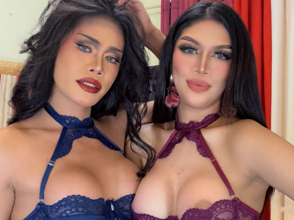 Dos mujeres de HottyBananaDollyz con senos grandes, vestidas con lencería de encaje, una en azul y otra en morado, de pie muy cerca, cabello oscuro, maquillaje intenso, lápiz labial rojo, mirando a la cámara..