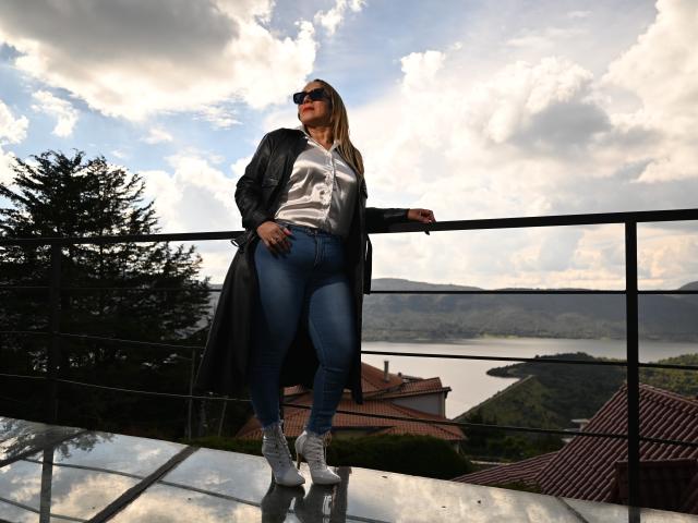 DianeOasis stands on a rooftop, wearing a black leather coat, white shirt, blue jeans, and white sneakers. She has blonde hair, sunglasses, and a hand on her ass.