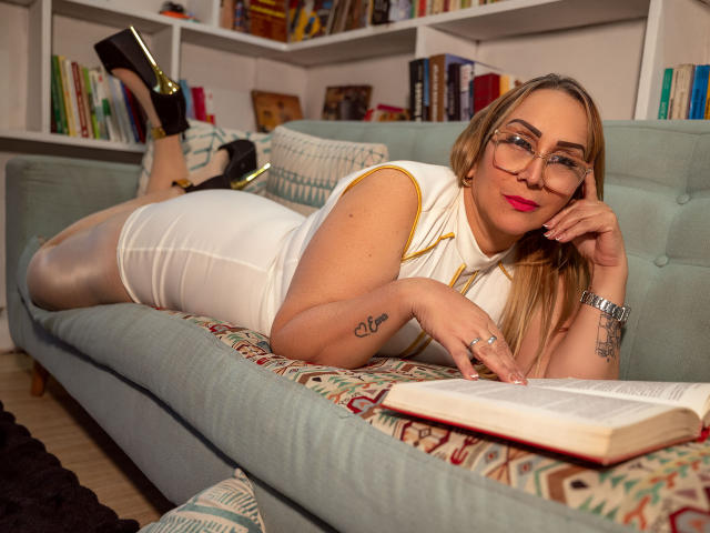 DianeOasis, recostada en un sofá azul, vestido blanco, gafas, tacones negros con detalles dorados, lápiz labial rojo, tatuaje en el brazo derecho, leyendo un libro, piernas levantadas, sexy, relajada..