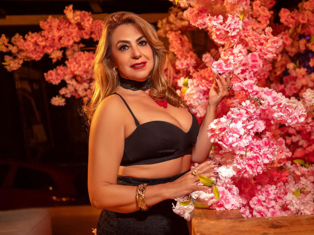 LylaGreen with blonde hair, black bra, and lace choker, touching pink flowers, wearing gold bracelet, smiling, showing her tits, in a night setting with pink blossoms.