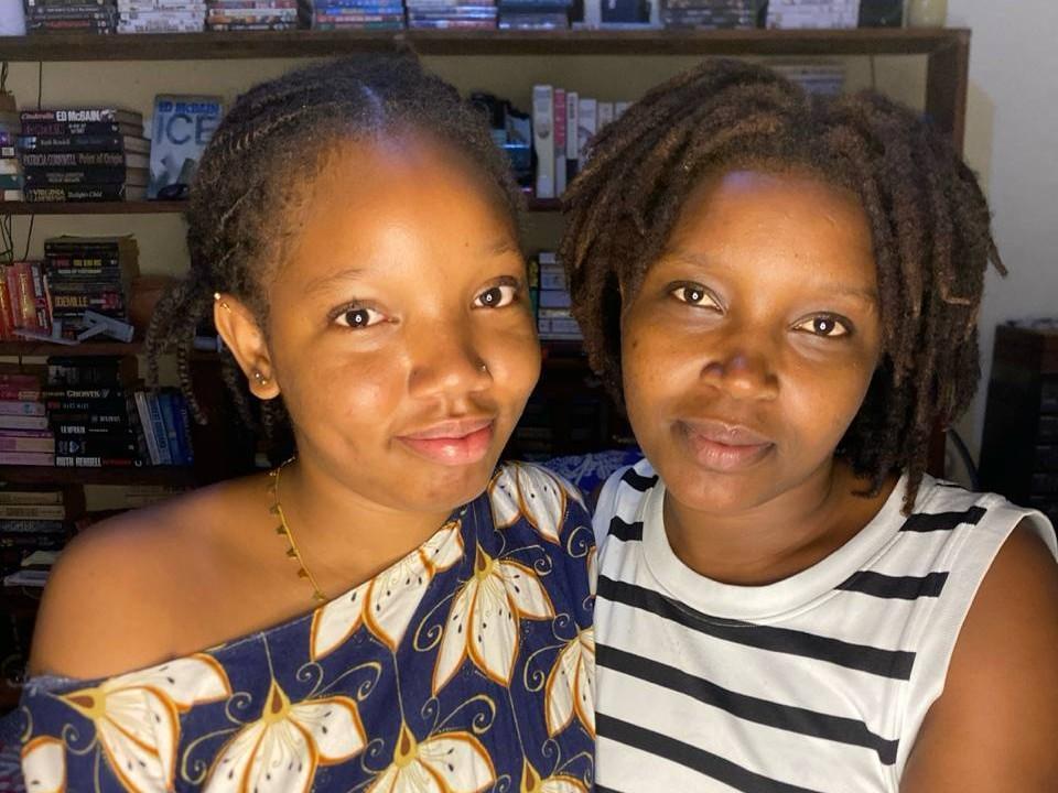 Deux SexyArya et deux Tiffy séduisantes, à la peau foncée, souriantes, debout côte à côte. L'une a les cheveux bouclés, porte une robe bleu marine avec des motifs jaunes et un collier en or. L'autre a des dreadlocks, et porte une chemise rayée blanche et noire..