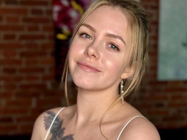 HannieBunnie-hot with blonde hair, green eyes, white spaghetti straps, subtle smile, tattoo on left shoulder, gold earrings, brick wall background.