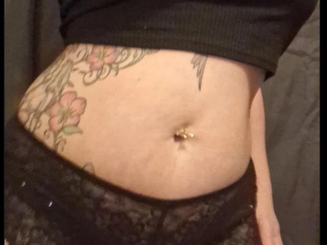 FrenchieLilieFleur's middel, met een piercing in de navel, een bloemen-tatoeage aan de rechterkant, en kanten ondergoed, waarbij een stukje van haar zwarte top te zien is..