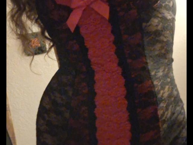 De kantjurk van FrenchieLilieFleur heeft een zwart-grijs bloemenpatroon met een roze, rozenachtige versiering in het midden, en is afgewerkt met een roze strik. Haar krullende bruine haar is zichtbaar..