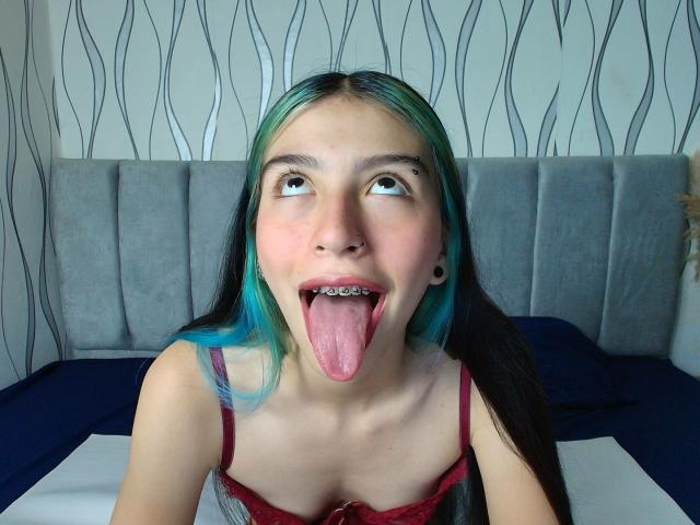 AbbyHillsI with blue-green hair, tongue out, showing metal teeth, wearing red lingerie, looking up, has black ear tunnels, pale skin, sitting on a bed with dark pillows.