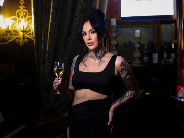 HottyNicole with tattoos, black cut-out dress, holding a glass of champagne, standing in a dimly lit room, looking at the camera with a slight smile.