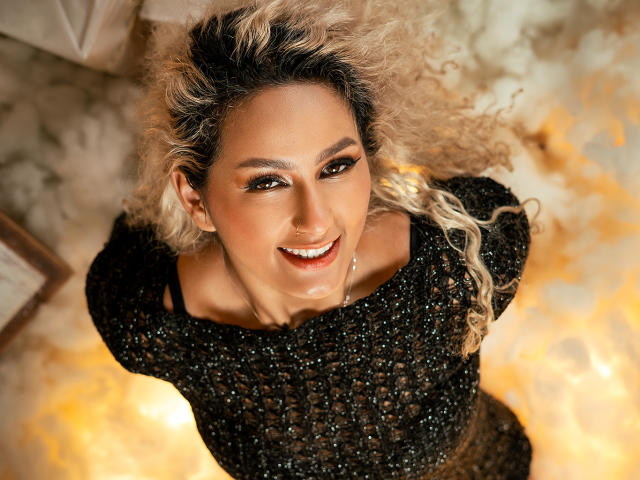 FloraSaenz with blonde curly hair, wearing a sparkly black top, smiling up at the camera, has a cute nose ring and glowing skin, on a warm, textured floor.