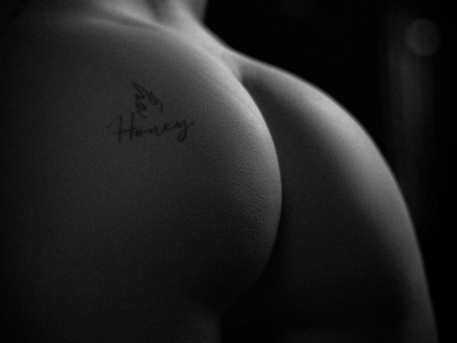 NyraVanel's butt is in the foreground, with a tattoo reading "Honey" on the right cheek. The black and white photo emphasizes the smooth, round texture of her skin.
