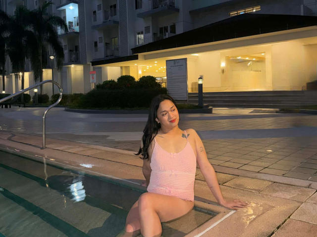 AmaraHugeCock, assise au bord d'une piscine, portant un maillot de bain rose, mouillé, une pièce, qui dévoile ses seins, avec un tatouage sur son bras droit, la nuit..