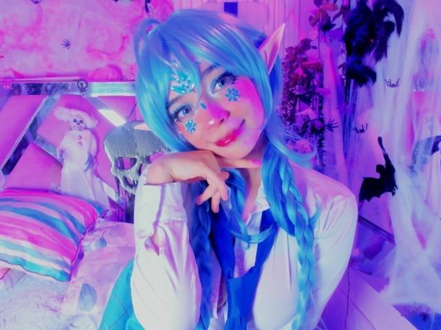KiraBerry with blue braided hair, elf ears, snowflake face paint, white shirt, blue vest, pink-purple room, smiling, resting chin on hand, fluffy white pillow, skull decoration.