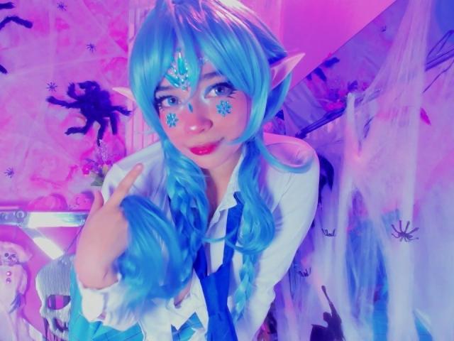 KiraBerry with blue elf ears, blue braided hair, white shirt, blue tie, snowflake makeup, pointing, pink-purple lighting, smiling. Her cute, fairy-like face, and elf-like features are prominent.