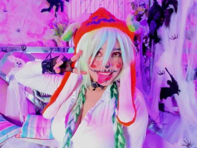 KiraBerry with colorful hair, zombie face paint, rainbow hat, white shirt, and fishnet glove, making a peace sign in a pink-lit room. Boobs, green braids, and a cute ass.